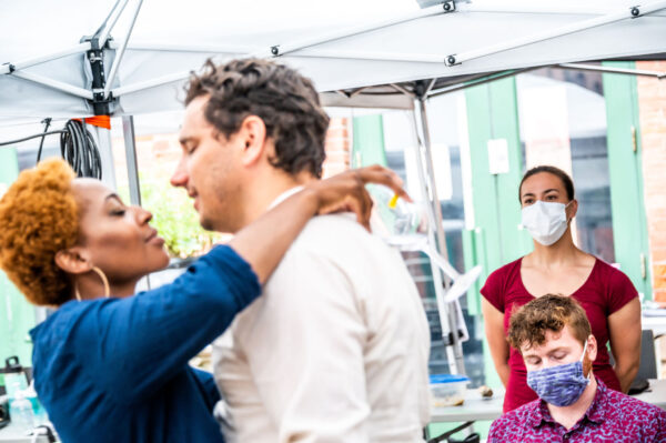 in the foreground a pair of actors rehearses the preparation for a kiss, observed by stage manager and intimacy choreographer in the background.